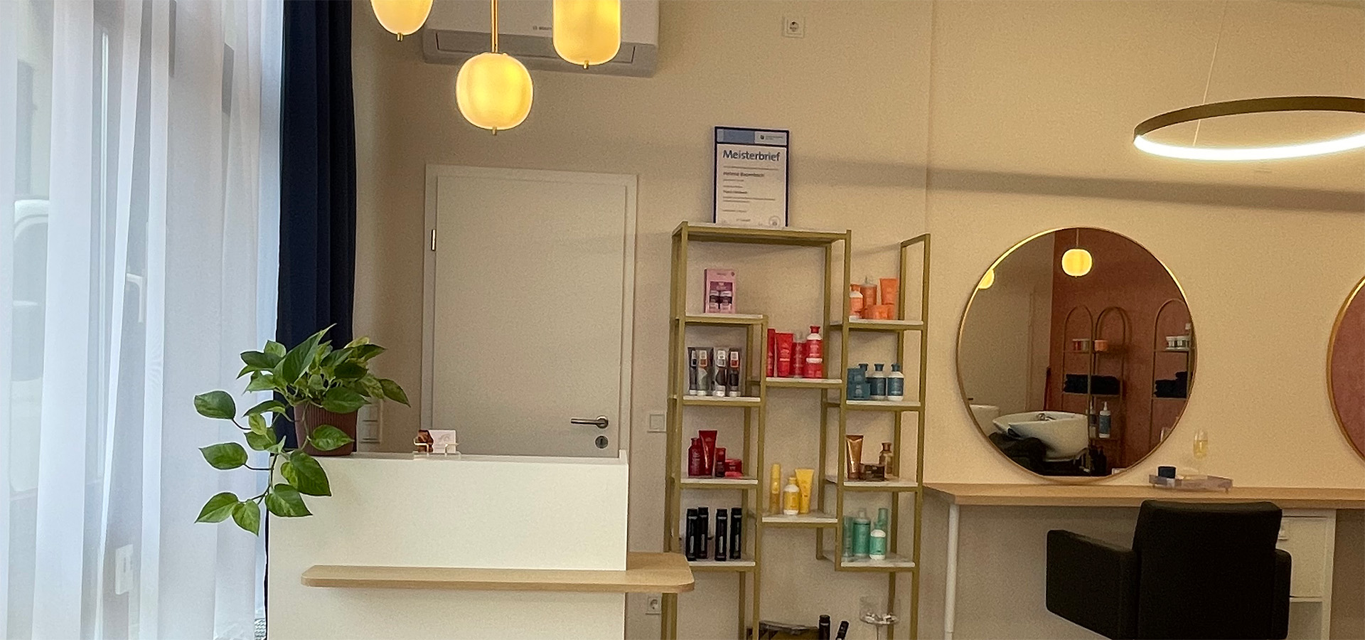 Modernes Friseursalon-Interieur mit Empfangstresen, Pflanze, Regal mit Haarpflegeprodukten, runden Spiegeln und Stylingstühlen – ruhiger, persönlicher Rahmen für hochwertige Haarservices von Helene Baumbach in Neuhofen.